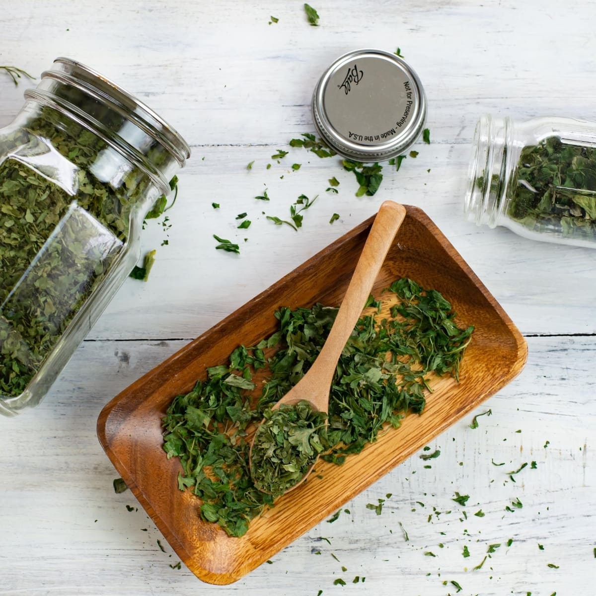 dehydrated parsley in a long wooden bowl with a small wooden spoon. In the background are two jars filled with dried parsley to show how to store the dried herb.