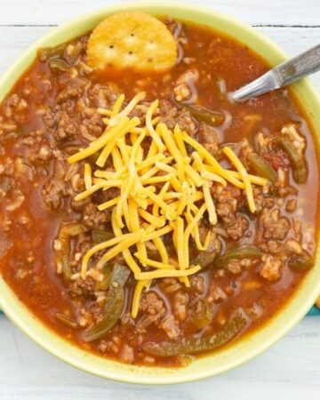 Overhead photo of a green bowl filled with Instant Pot Stuffed Pepper Soup on a cloth napkin, a spoon and cracker in the bowl, topped with shredded cheddar cheese