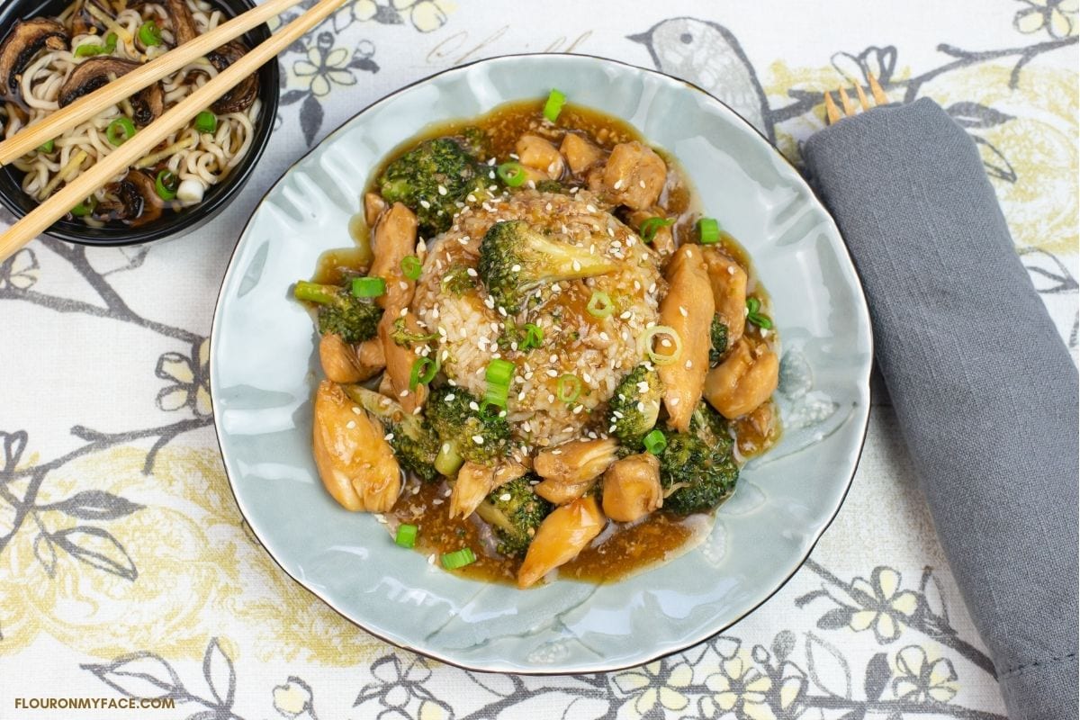 Chicken and Broccoli served over white rice on a grey glass plate with a bowl of soup in the background and a grey cloth napkin next to the plate