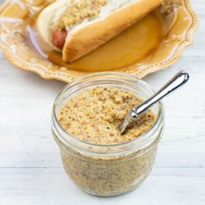 Homemade Grainy Dijon Mustard in a glass jar with a plate in the background with a hot dog and sauerkraut spread with grainy Dijon Mustard