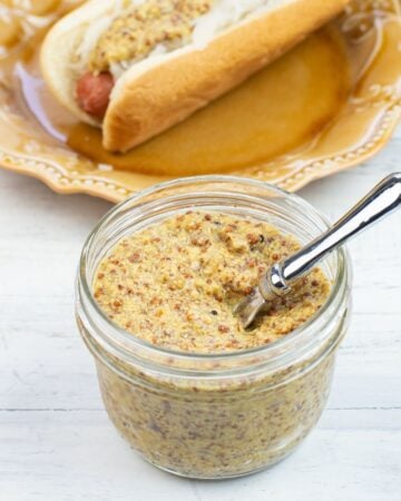 Homemade Grainy Dijon Mustard in a glass jar with a plate in the background with a hot dog and sauerkraut spread with grainy Dijon Mustard