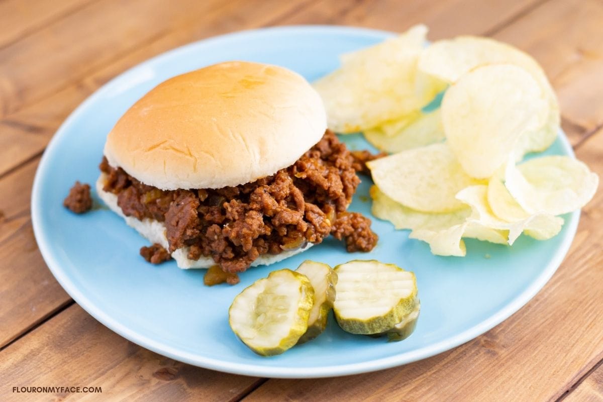 Sloppy Joes served on a bun, with, potato chips and sliced pickles on a plate.