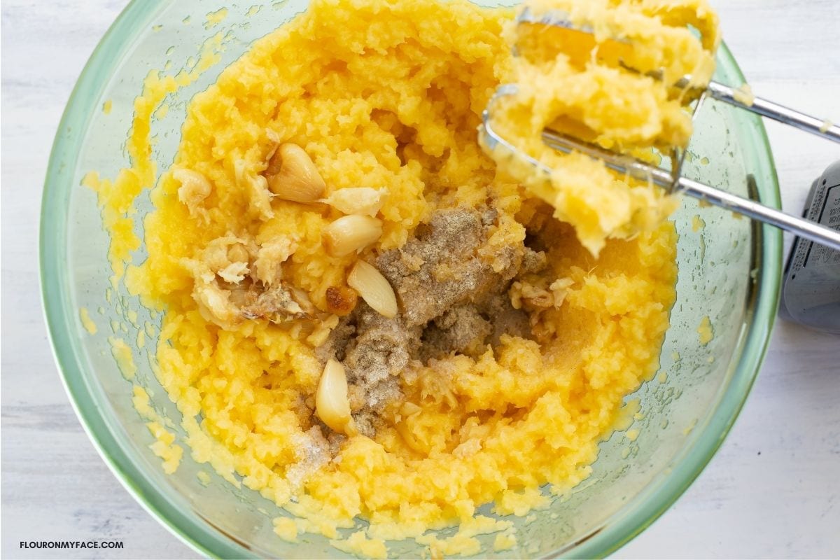 Adding roasted garlic and spices to mashed rutabaga in a mixing bowl.