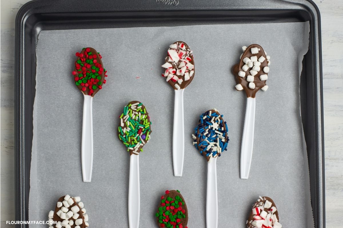 chocolate dipped spoons decorated with holiday sprinkles on a cookie sheet.