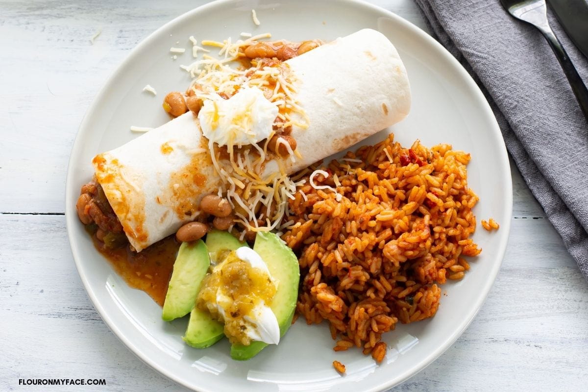 Chicken burrito served with rice and sliced avocado on a plate.