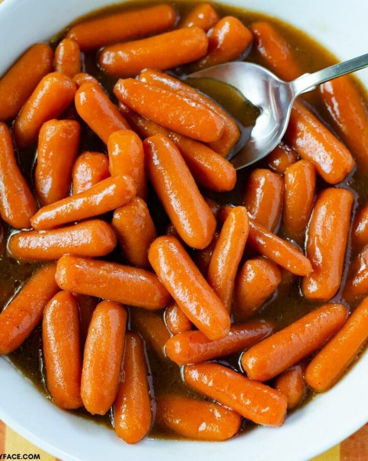 A white bowl filled with glazed carrots with a serving spoon.