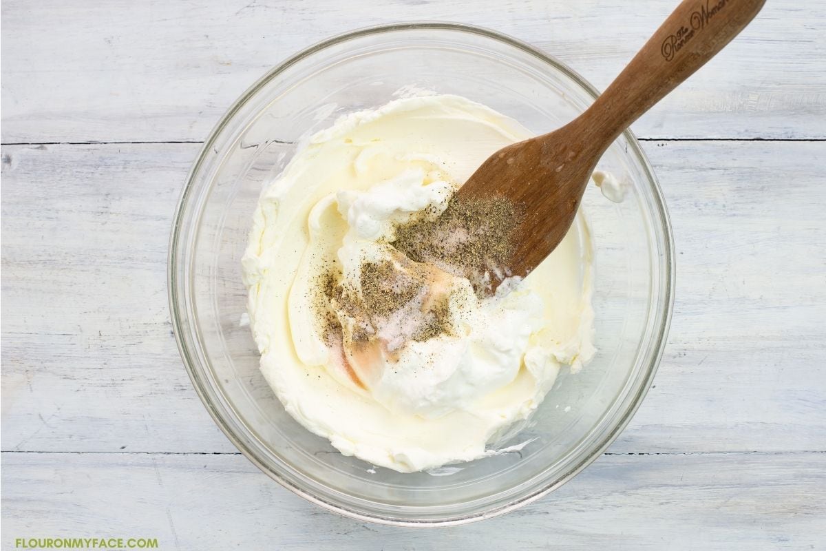 Mixing softened cream cheese, sour cream, salt and pepper together in a bowl with a wooden spoon.