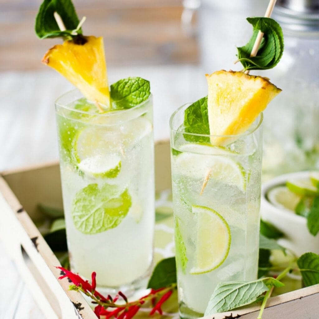 2 tall glasses filled with pineapple sage mojito, garnished with pineapple wedge and lime wedges.