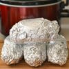 Four baking potatoes wrapped in aluminum on a cutting board in front of a crock pot.