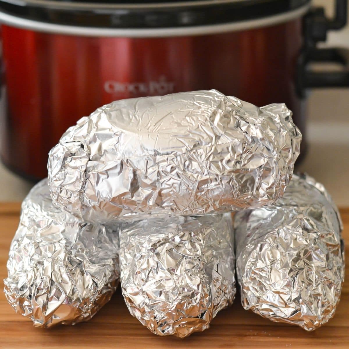 Four baking potatoes wrapped in aluminum on a cutting board in front of a crock pot.