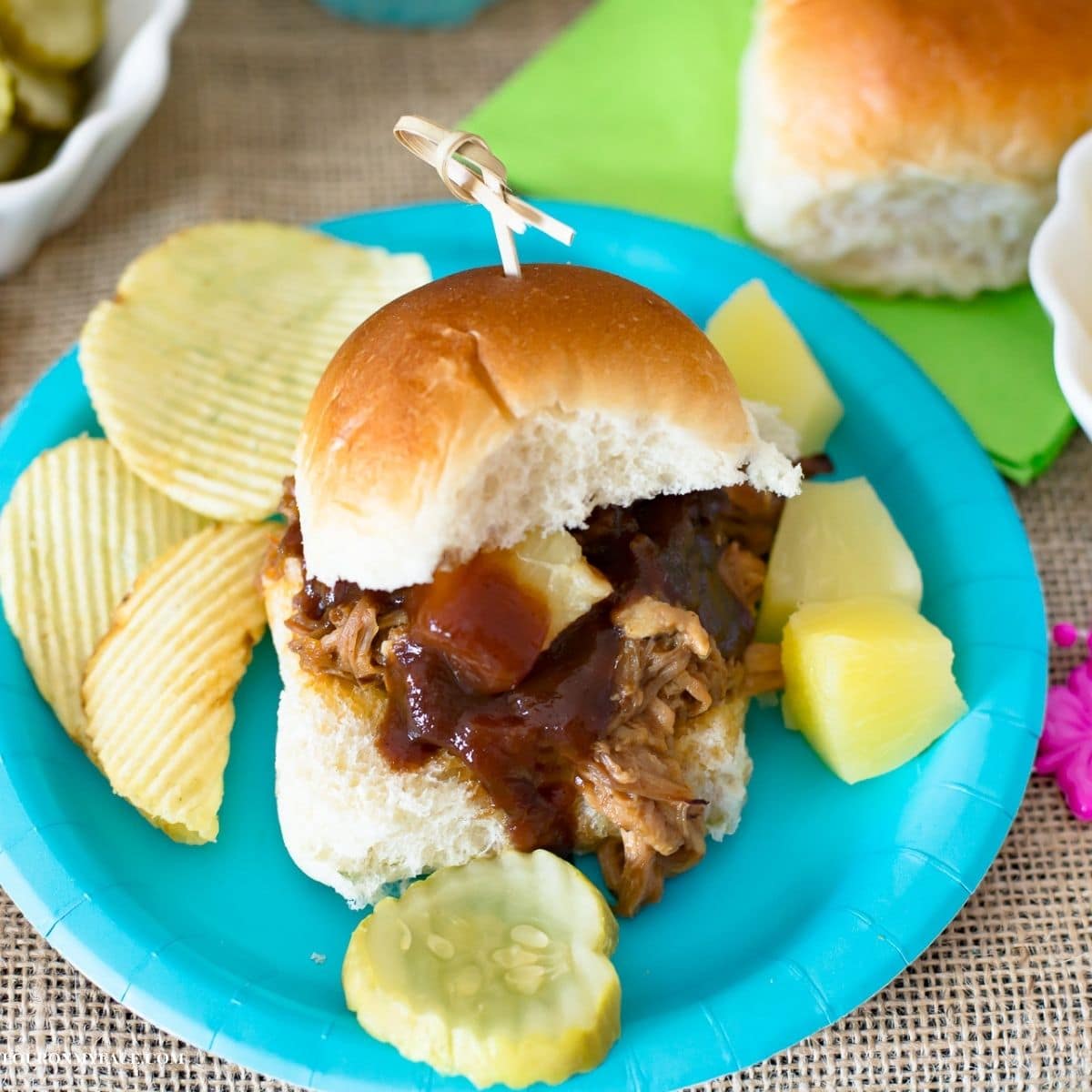 A pineapple pulled pork slider with chips and pineapple chunks on a plate.