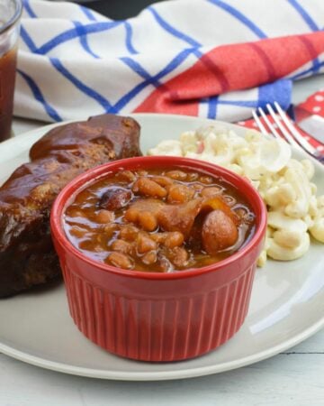 A small bowl filled with baked beans on a plate with ribs.