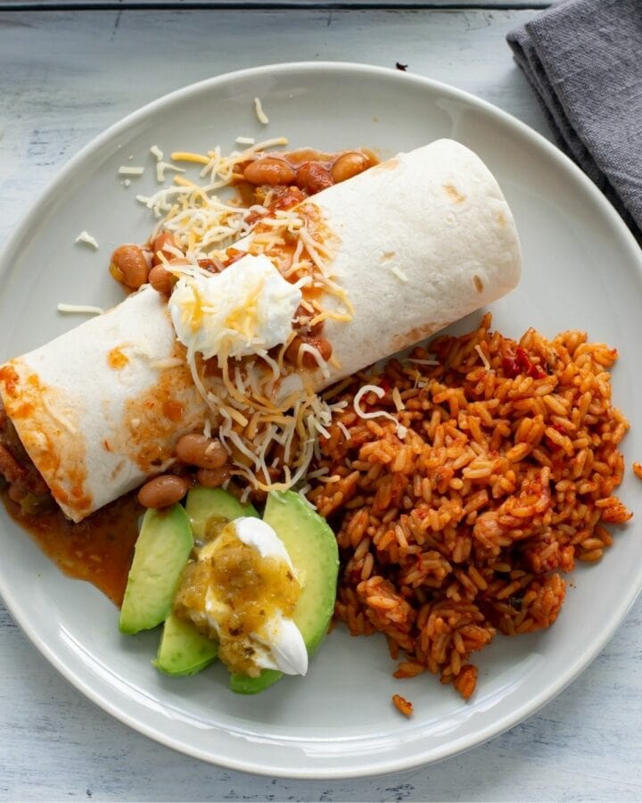 A dinner plate with crock pot chicken and bean burritos, sliced avocado and Spanish rice.
