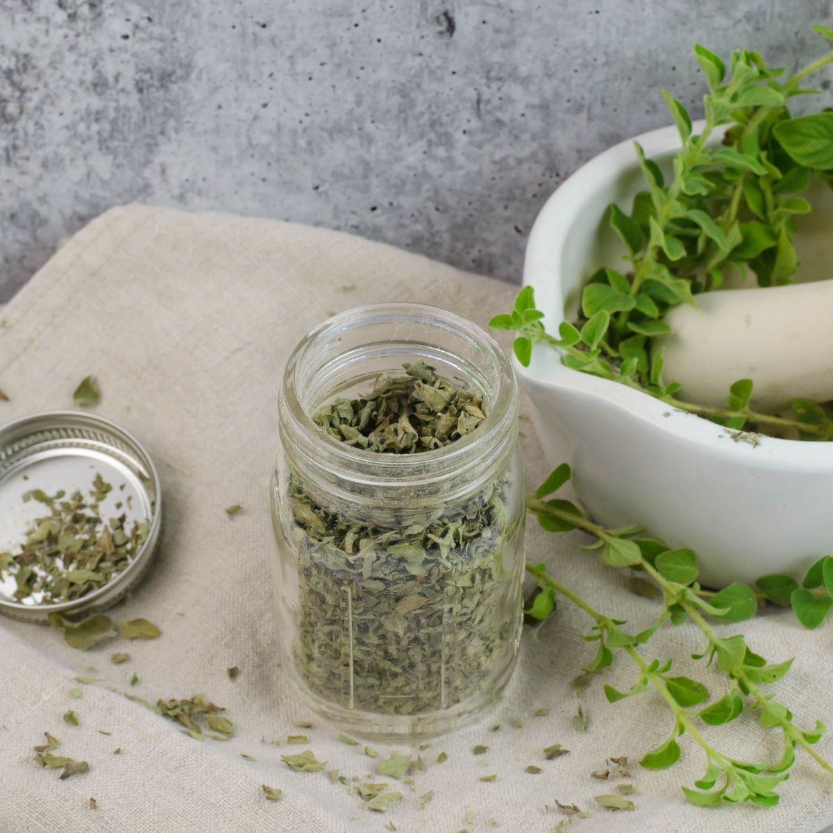 Overhead photo of dehydrated oregano in a spice bottle on a napkin with a white mortar and pestle in the background.