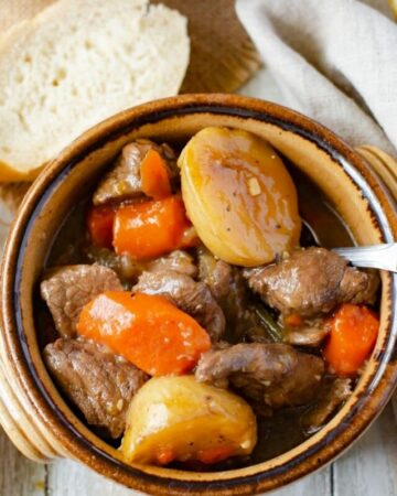 A brown soup crock filled with beef stew with chunks of potatoes, carrots and a thick beef gravy.