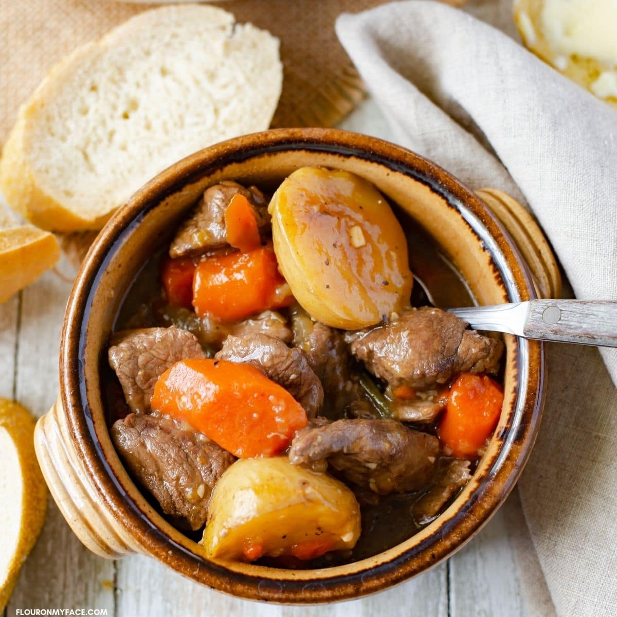 Overhead image of a crock like bowl filled with beef stew with sliced french bread around the bowl.