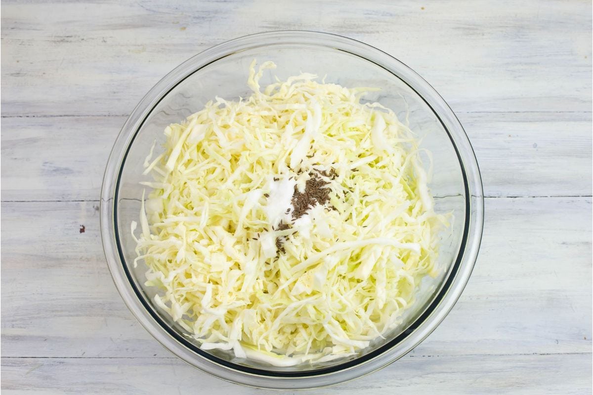A large glass bowl filled with shredded cabbage, with a tablespoon of salt and a teaspoon of caraway seed in the center.