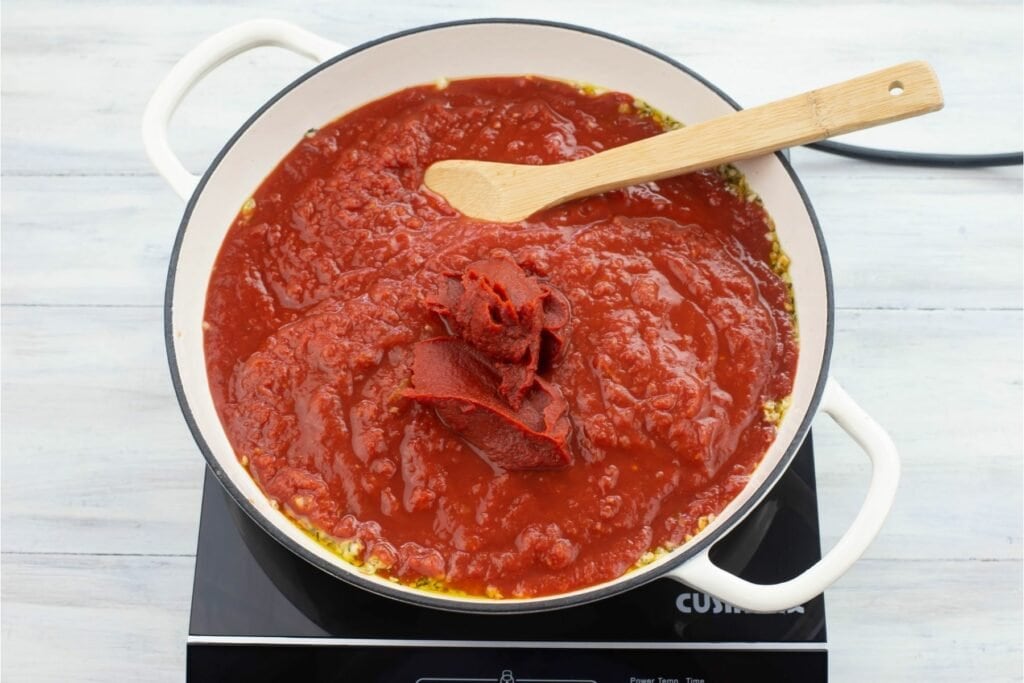 Combining crushed tomatoes, tomato paste, sauteed garlic and rosemary in a pan.