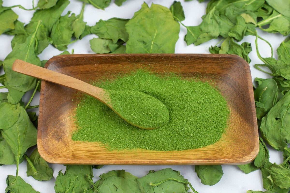 A pile of homemade spinach powder in a wooden bowl with a scoop on a wooden spoon.