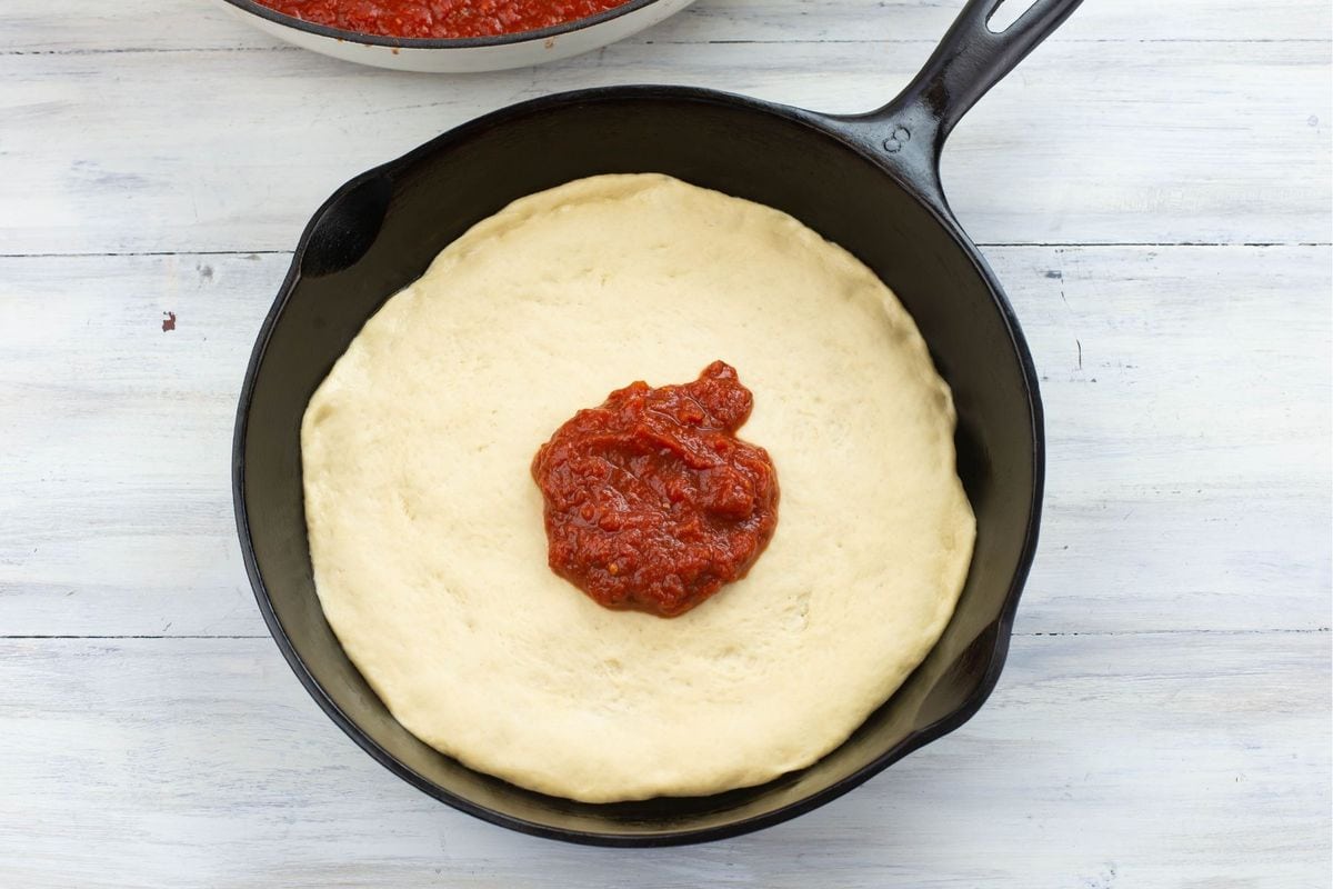 Cast iron skillet with uncooked pizza dough and pizza sauce.