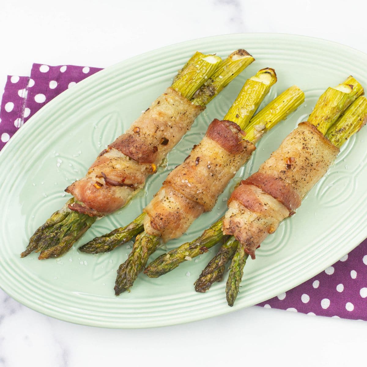 Three bacon wrapped asparagus bundles on a pale green serving plate.