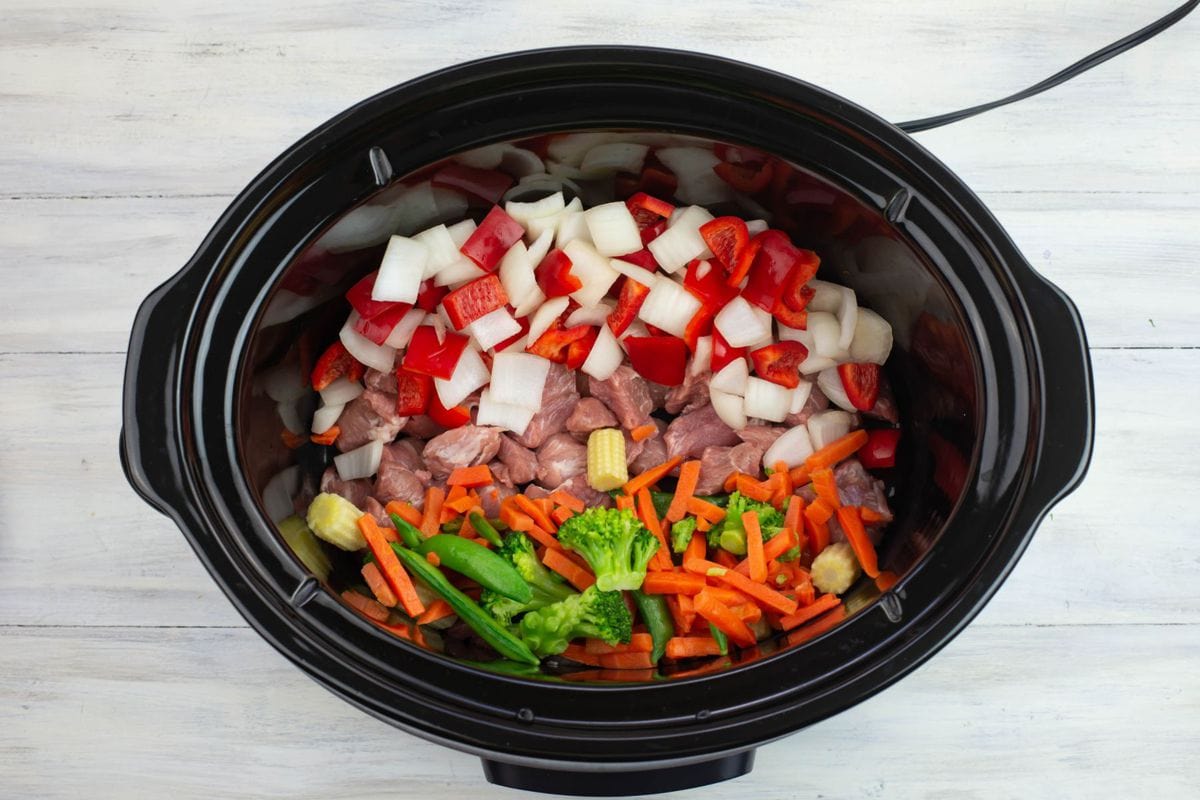 Cubed pork and prepared vegetables in the bottom of a slow cooker.