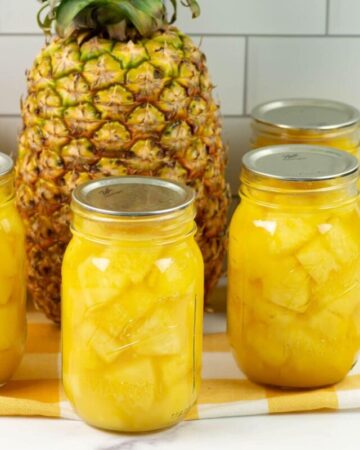 Pint canning jars filled with canned pineapple chunks with a whole ripe pineapple in the background.