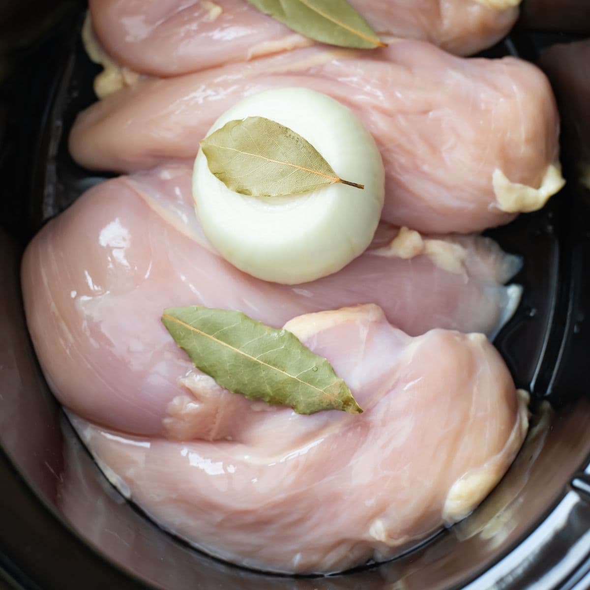 Boneless chicken breasts with and onion and dried bay leaves before cooking in a slow cooker.