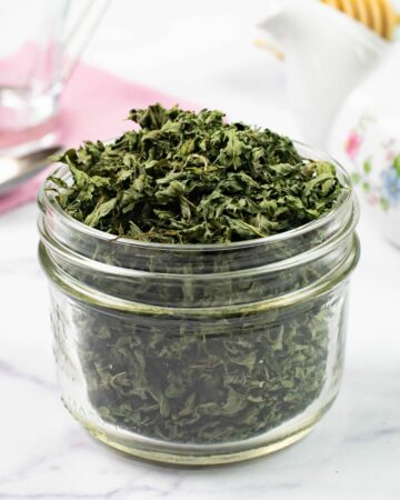 A small glass jar filled to overflowing with dried mint, a tea pot, spoon and napkin in background.
