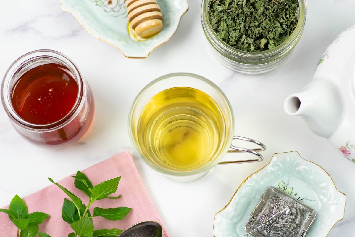 Overhead image looking down at a tea cup filled with mint tea with mint, honey, a teapot and tea ball on a tabletop.