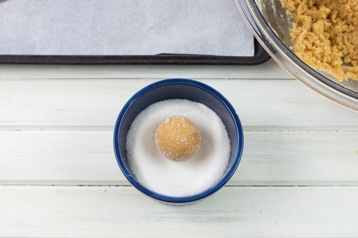 Dipping cookie dough balls into granulated sugar in a small bowl.