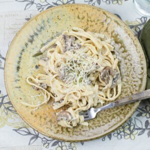 Mushroom Alfredo with milk served over cooked fettucinne noodles on a brown clay dinner plate.