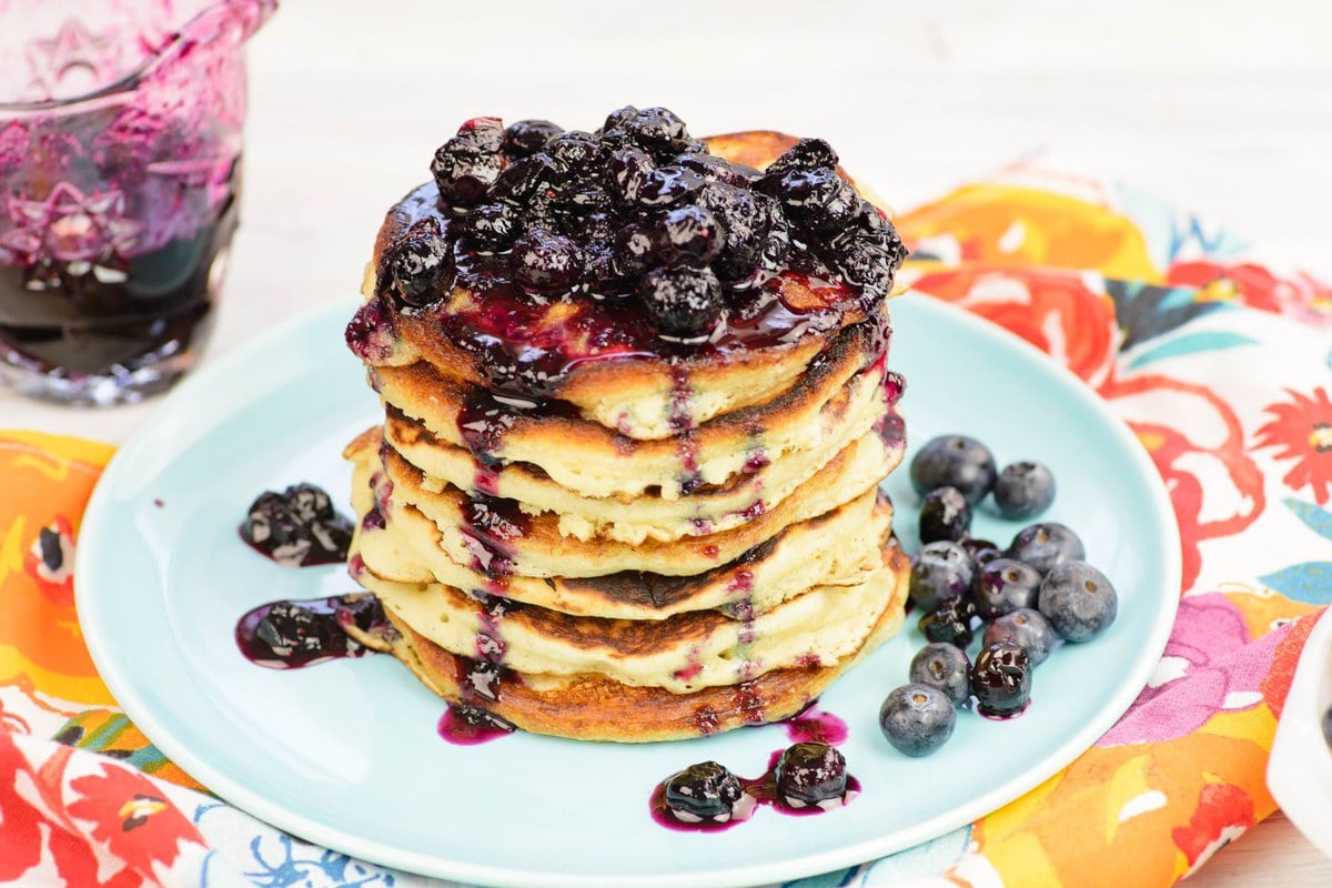 A stack of pancakes on a plate with blueberry sauce drizzled over them.