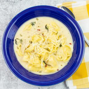 Chicken Tortellini Alfredo Soup in a blue bowl with a cloth napkin and spoon.
