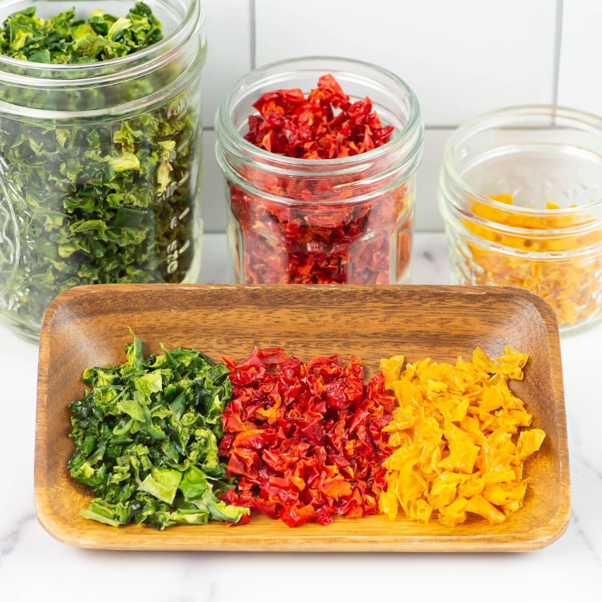 Green, red, and yellow dehydrated bell peppers in a wooden bowl with filled jars in the background.
