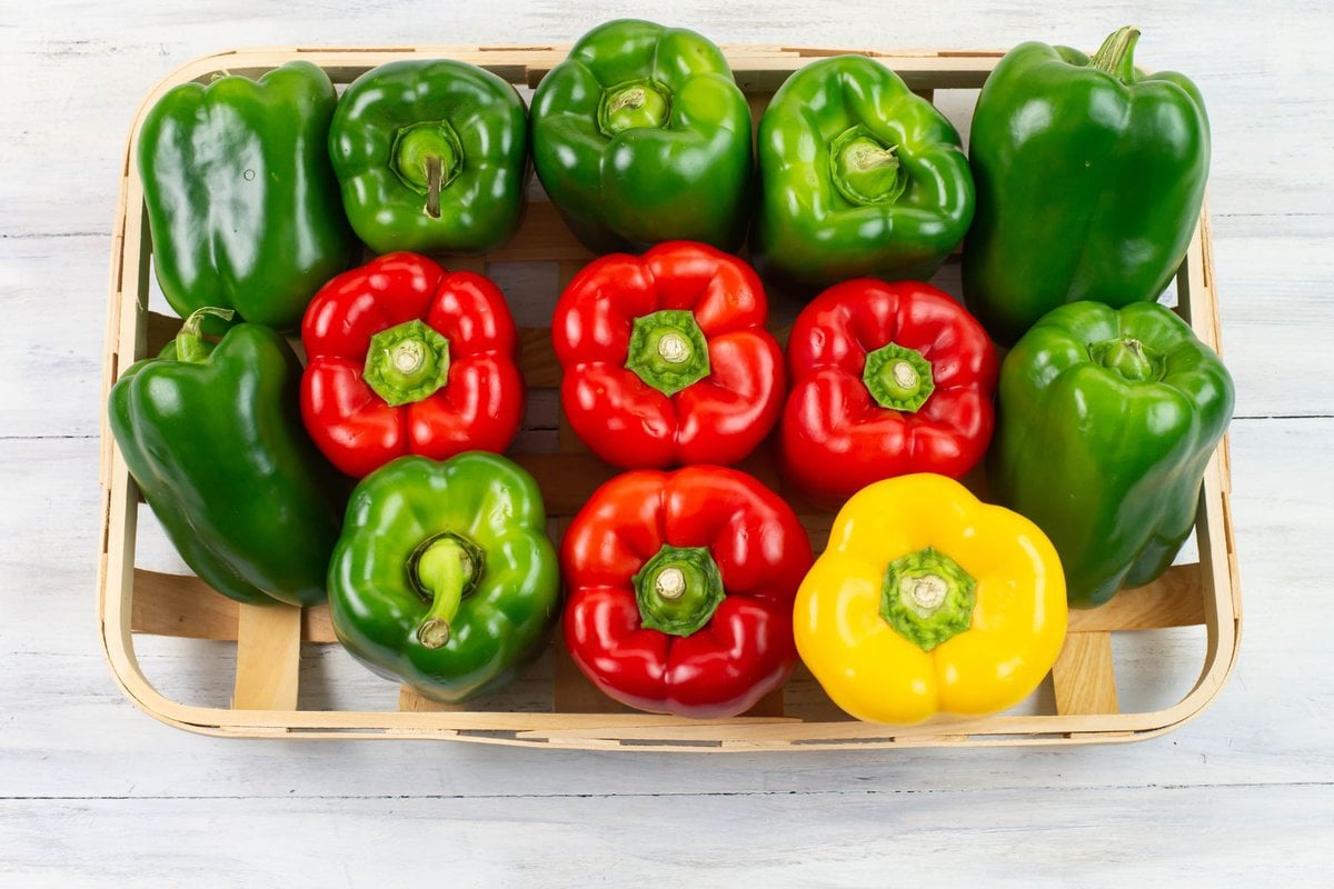 Ripe green, red and yellow bell peppers in a flat wooden basket.