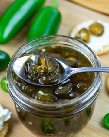 A spoonful of Candied Jalapenos resting on the edge of a mason jar rim with crackers and cream cheese in the background.