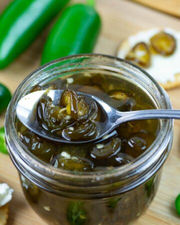 A spoonful of Candied Jalapenos resting on the edge of a mason jar rim with crackers and cream cheese in the background.