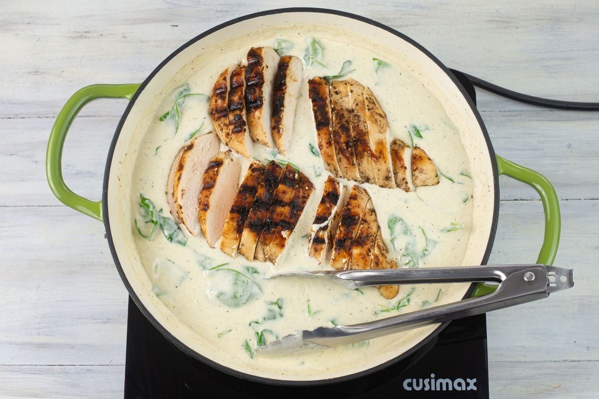 Adding sliced grilled chicken to a skillet of hot spinach alfredo sauce.