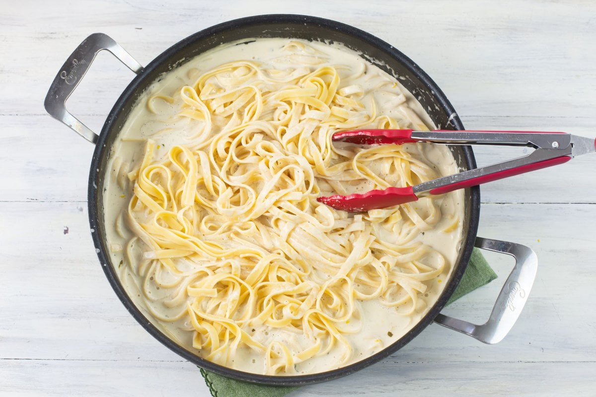 Tossing fettuccine pasta noodles with creamy lemon garlic alfredo sauce in a skillet.