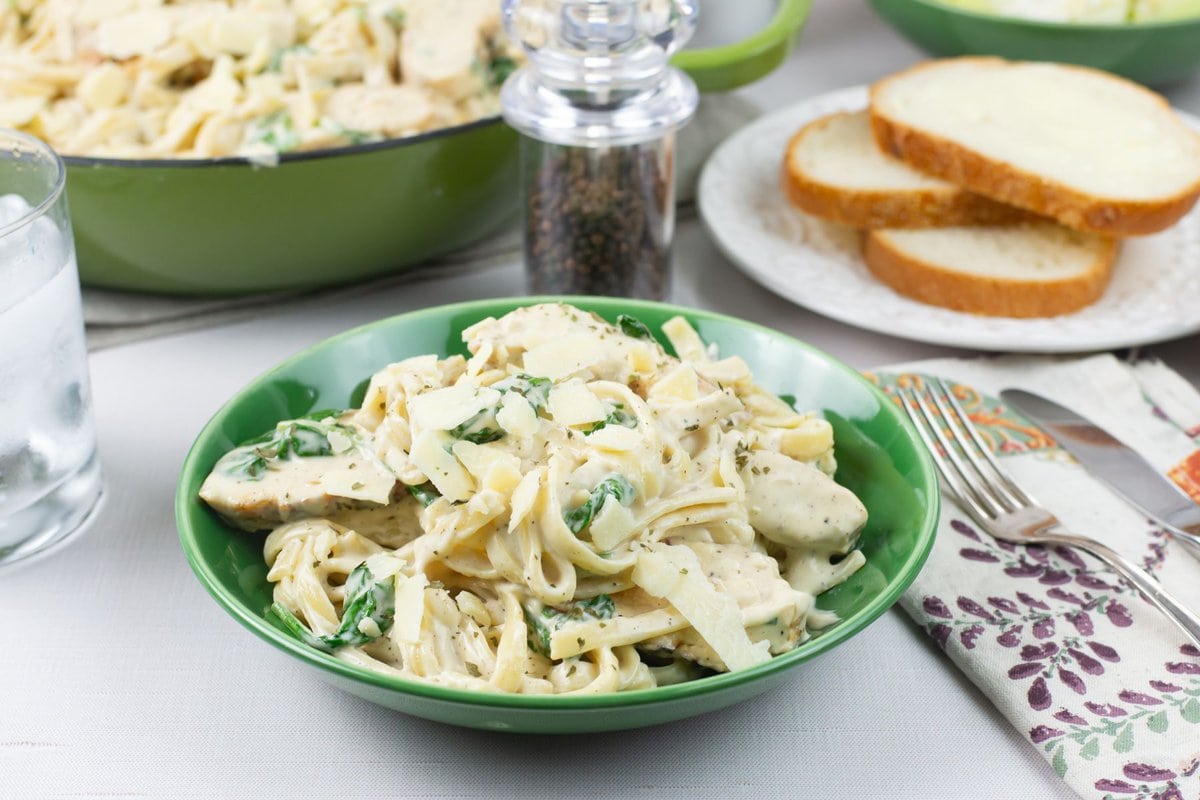 A bowl of creamy chicken spinach fettuccine alfredo served with bread and a salad.