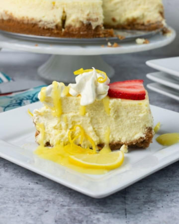 A slice of Lemon Cheesecake on a plate garnished with whipped cream,. lemon curd, and a slice of strawberry.