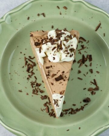 A thin slice of Nutella cheesecake on a green dessert dish, garnished with whipped cream and chocolate curls.