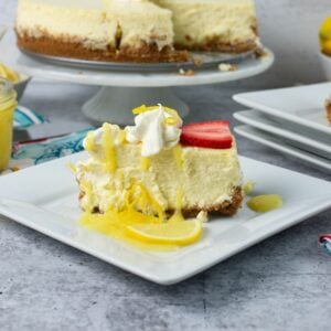 A slice of Lemon Cheesecake on a plate garnished with whipped cream,. lemon curd, and a slice of strawberry.