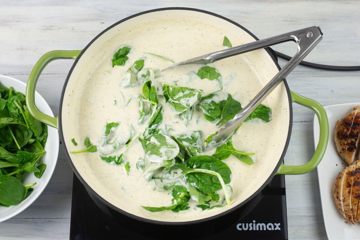Adding fresh raw baby spinach leaves to a skillet of hot alfredo sauce.