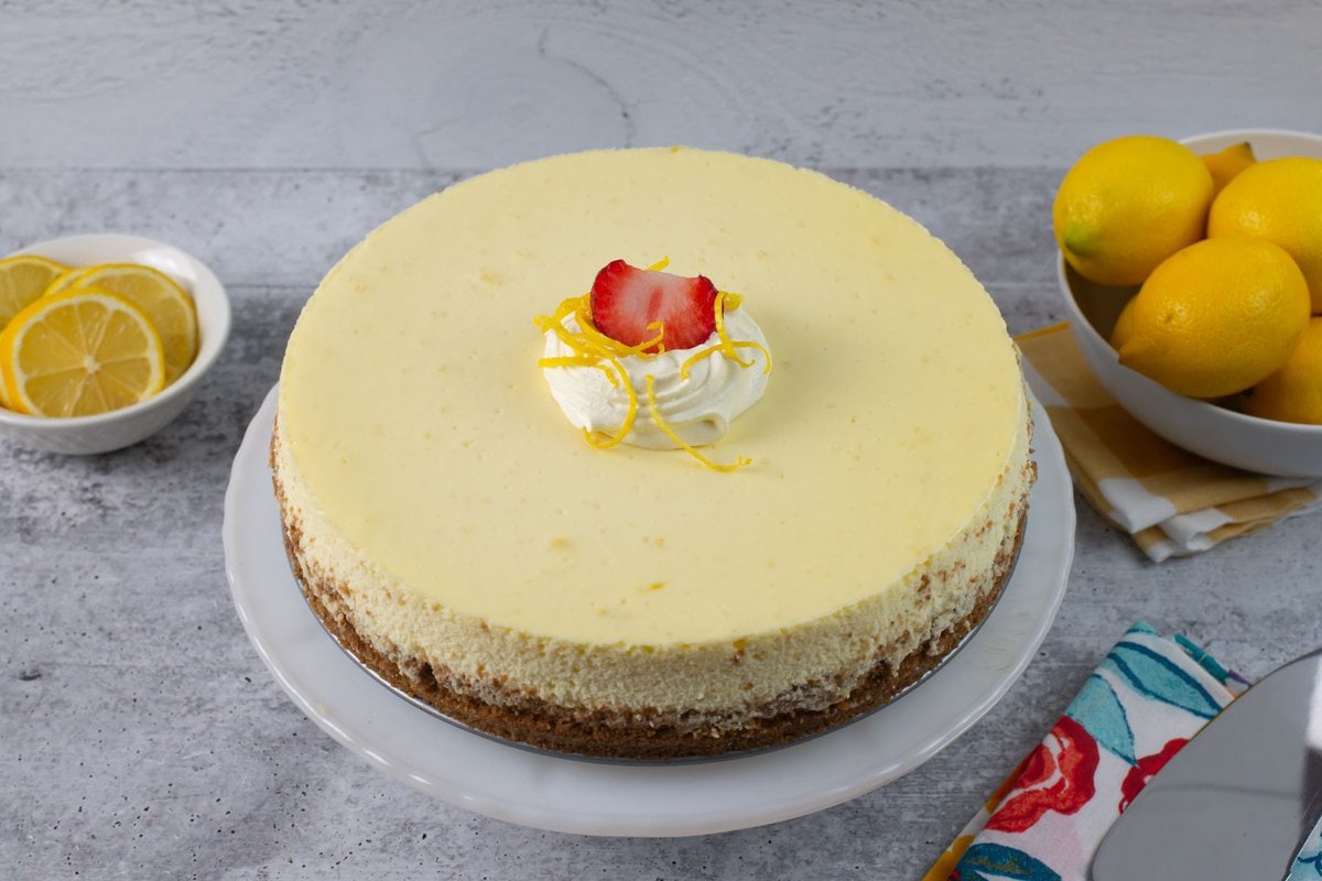 a 9-inch uncut Lemon Cheesecake garnished with whipped cream and sliced strawberry on a white cake stand.