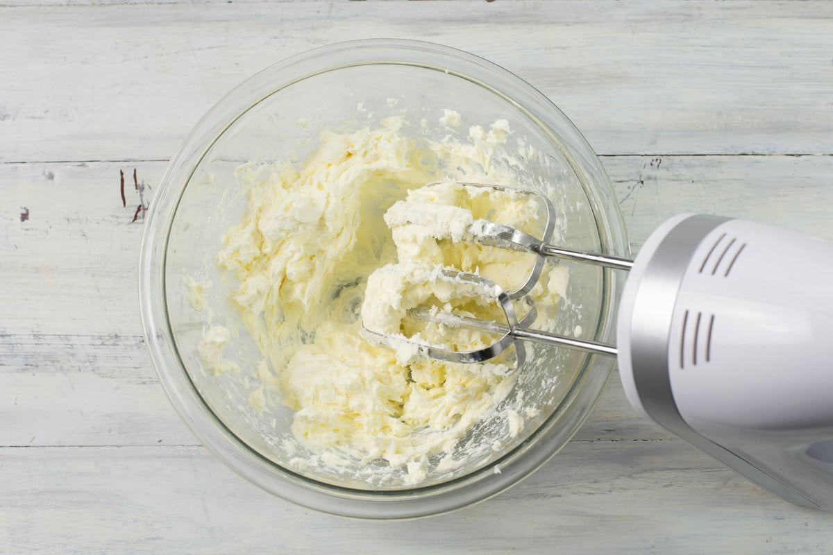 Beating cream cheese with a hand mixer in a glass bowl.