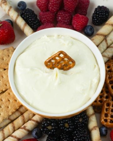 Closeup image of a bowl filled with cheesecake dip with, berries, crackers and pretzels surrounding the bowl.