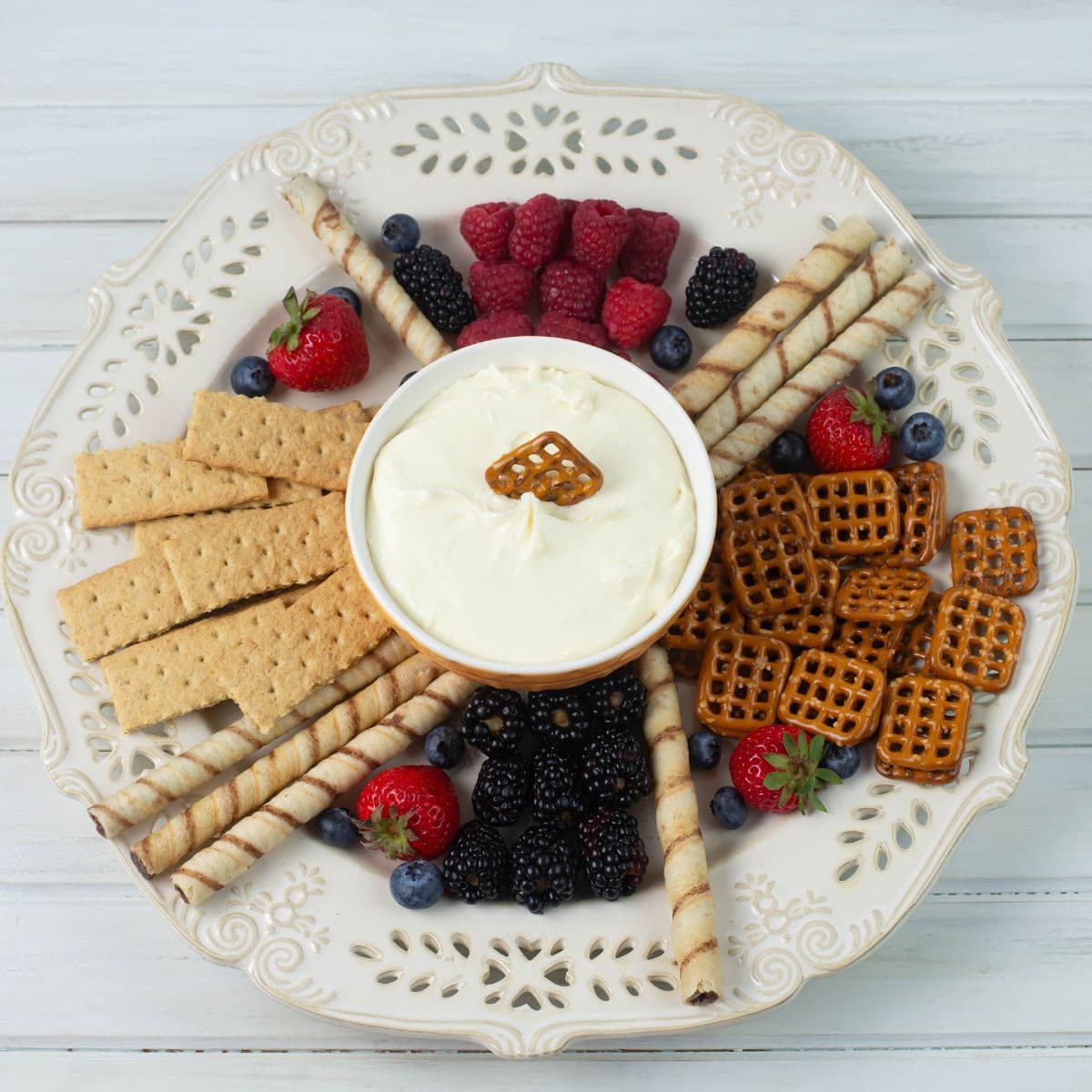 A large platter with a bowl of cheesecake dip in the center surrounded by fruits, cookies and pretzels.