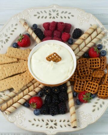 A large platter with a bowl of cheesecake dip in the center surrounded by fruits, cookies and pretzels.